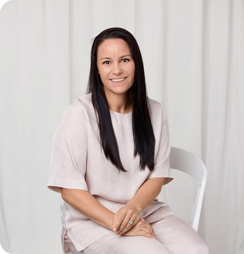 Smiling woman seated on a white chair with a notebook in a minimalist studio.