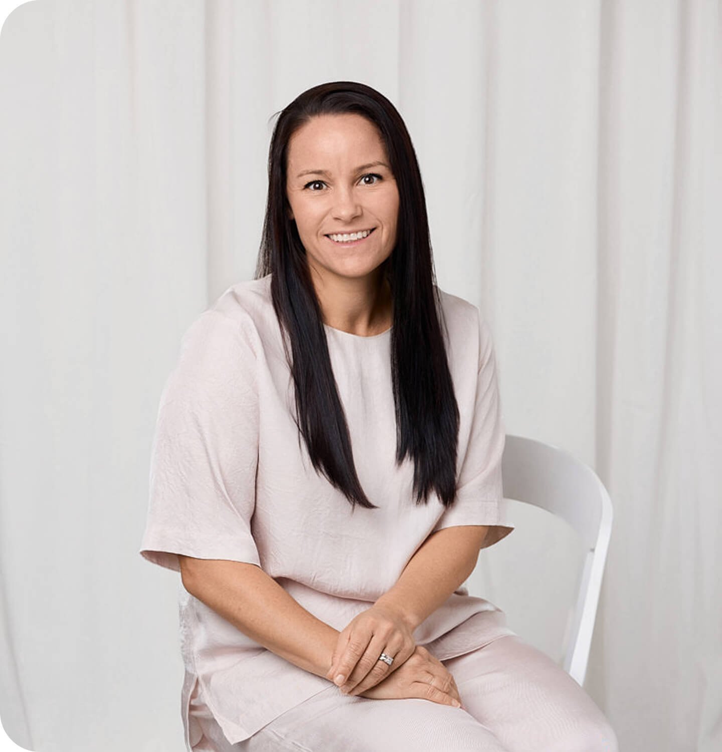 Smiling woman seated on a white chair with a notebook in a minimalist studio.