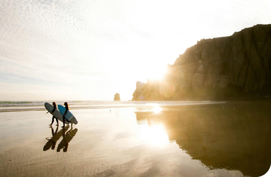 Two surfers walking along a sunlit beach at low tide.