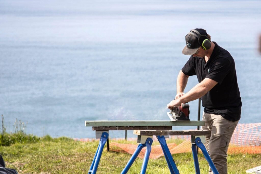 Builder cutting a board on sawhorses beside the ocean.
