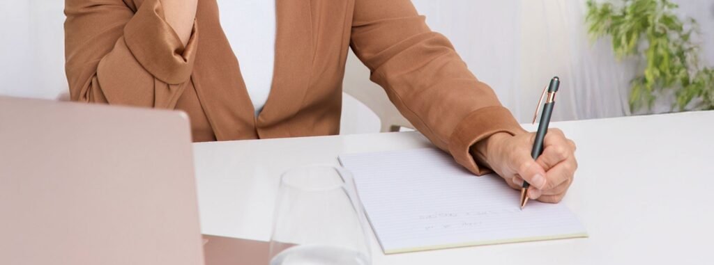 Person in a tan blazer writing notes at a white desk with a laptop.