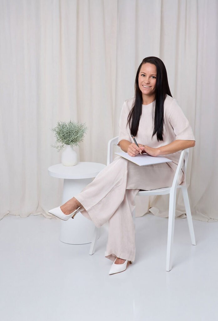 Smiling woman seated on a white chair with a notebook in a minimalist studio.